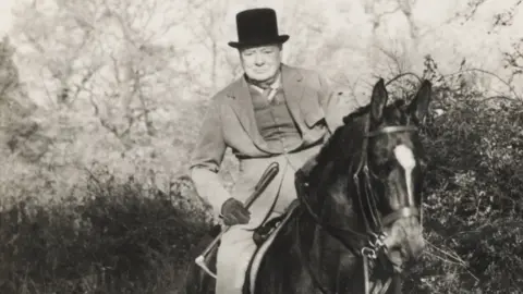 Dominic Winter Auctioneers Black and white image of Sir Winston Churchill on horseback. He is well dressed and is wearing his iconic black hat.