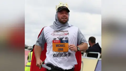 Michael Donkin Michael running wearing a knight's costume with a red cape and carrying a plastic sword. His name is on his T-shirt