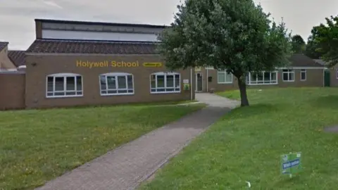 Holywell School in Bedfordshire as seen on Google street view. It has a yellow sign outside and its name in yellow text on the building. A path leads to the school entrance through a grassy area.