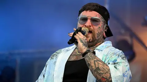 Getty Images Singer Teddy Swims holding a microphone to his mouth. He is wearing sunglasses, a black cap, a black top and an unbuttoned blue shirt. He has tattoos on his arms and face. 