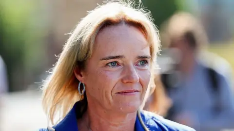 A woman with blond hair and wearing a blue suit, walking outside. Other people - out of focus - are in the background