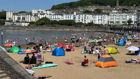 Many people are sitting on sand by a large marine lake in Weston-super-Mare.