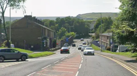 Google streetview of Grane Road, with cars on the carriageway. Terraced houses line the left of the road and trees on the right. Green hills can be seen in the backdrop.