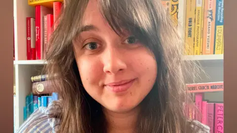 A young woman with long brown hair, wearing a blue and white striped shirt, smiles at the camera in front of a shelf lined with books. 