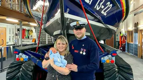 Bridlington RNLI A man in an RNLI navy uniform poses for a photo with his wife and newborn son, in front of a blue lifeboat.