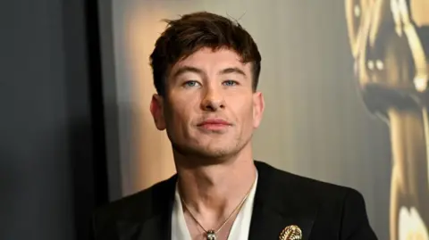 Getty Images Barry Keoghan standing in front of a black and gold bankground. He is wearing a while shirt and black blazer