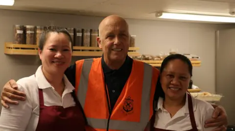 Stella Maris Paul Atkinson wearing a high-vis jacket standing in a kitchen with his arms around two women in aprons. 