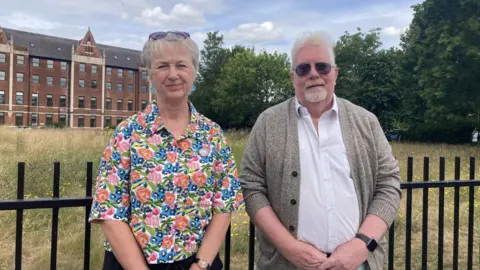 A woman in a flowery shirt and a man in a shirt and cardigan are standing side by side in front of an inner city field that is overgrown with wild flowers and grass.