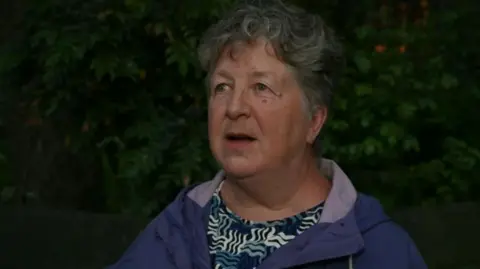 A woman with short grey hair stands in front of a hedge. She is wearing a blue top with a white, grey and black print, and a purple coat.