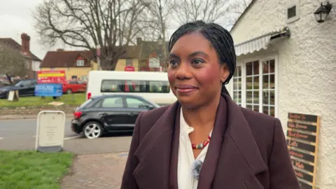 A woman wearing a dark purple suit and white shirt is standing outside a shop.