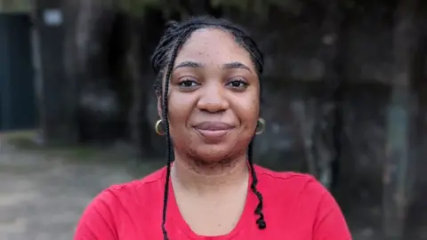 Portrait of Medlyn looking at the camera with a smile on her face. She has long braided afro hair, gold earrings and a red, round necked t-shirt. She is pictured outside, with a concrete wall behind her. 