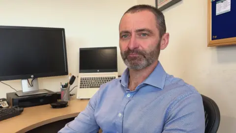 Picture of Tobin Cook looking at the camera while sat at a desk with a computer on it. He has short hair and a beard, and is wearing a blue button-up shirt.