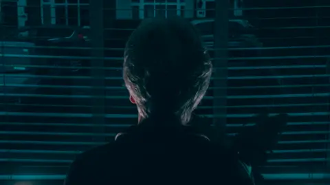 A woman stands looking out of a window. She has short hair and her back to the camera, to protect her identity. 