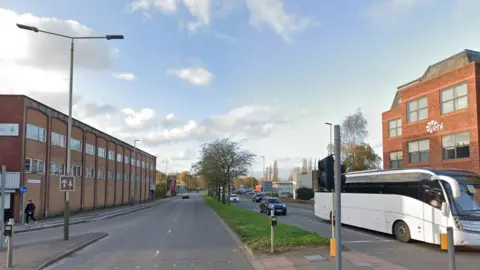 Google A dual carriageway road with trees along a grass central reservation. A white bus travels along the right-hand side of the image. The road is lined by office and industrial properties.