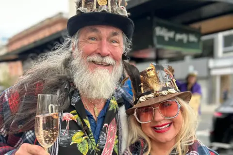 Melody and John Hennessee in Belfast smiling and holding glasses of prosecco. They are wearing cowboy hats and fun glasses.