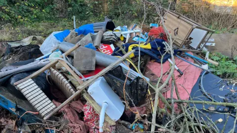 Domestic items, such as a toilet, wooden chair, bin, clothing and a rug, fly-tipped in a rural setting 