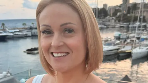 Jo Wallace A woman with bobbed blonde hair . She is smiling and looking at the camera and is stood in front of a slightly blurry background of a marina with lots of boats on the water and palm trees and buildings behind. 