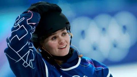 Maisie Hill looks on after her run in the first round in the Women's Snowboard Big Air qualification of the snowboarding competitions at the 2026 Winter Olympic Games, in Livigno, Italy. She is wearing a black helmet and blue snow gear with white pattern on it. She has one hand on her head.