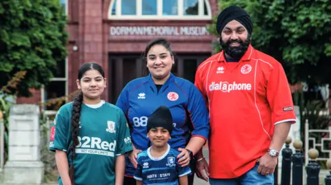 Tom Banks and Paul Thompson Jas, Anita, Aran and Tony are in a variety of Middlesbrough shirts, one green, two different blue designs and a red shirt. They are standing in front of the Dorman Memorial Museum with the two males wearing black turban head coverings. They are all smiling.
