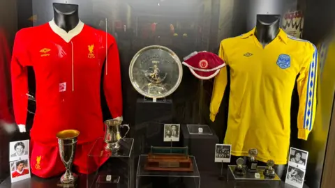 A red Liverpool kit and a yellow Chester City kit stand either side of a number of trophies won by Ian Rush