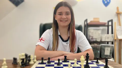 A white teenage girl with long light-brown hair with red streaks sitting at a table with a blue and white chess board and white and black chess pieces on it