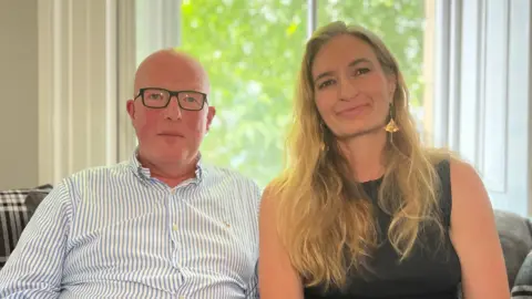 Andrew Woodward and Adele Denton are seated side by side on a grey sofa in front of a large window. Andrew wears a blue-and-white striped button-down shirt and glasses, while Adele wears a sleeveless black top and gold earrings. Natural light from the window illuminates the room, with green foliage visible outside and plaid cushions on the sofa.