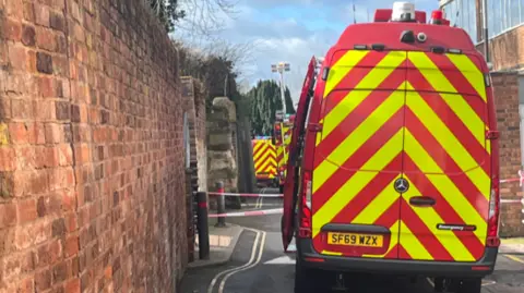 BBC A row of ambulances and fire engines can be seen in an alleyway. There is a long and quite high brick wall either side of the vehicles.