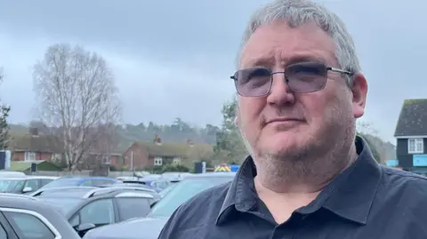 A man with dark tinted glasses and a blue shirt standing in a car park. He has grey hair and stubble. There are cars in the background and some small businesses.