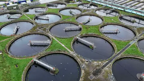 A general shot of a water treatment plant. There are several circular vats of water, each with an arm stretching from the middle to the edge of the circle. In between the circles are patches of grass.  