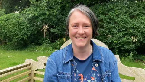 A woman is wearing a blue jacket and sitting on a park bench. She is smiling at the camera