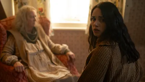 Daniel Alexander An elderly woman sat in a red arm chair looking into the distance. She has messy white hair and wears a house dress underneath an oversized cardigan. A younger woman sits opposite her, with shoulder length brown hair and a grey cardigan. She has a scared look on her face.