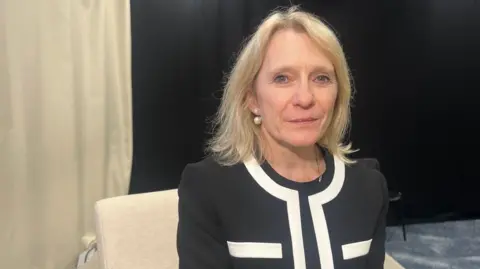 BBC Deputy Millar is smiling and is sitting in a beige chair in front of a black backdrop. She is wearing a black blazer with a white trim. She has blonde hair and is wearing pearl earrings. 