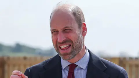 Prince William with his mouth wide open during a visit to Cornwall. He is wearing a blazer, blue and white stripey shirt and a burgundy tie. He has a trimmed beard and is balding on top of his head.