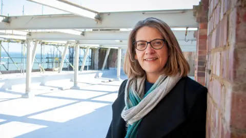 Dr Jess Steele - wearing glasses, a black coat as well as a blue and white scarf - stands on the roof a building. Steel girders and scaffolding can be seen behind her with the sea observable in the distance.