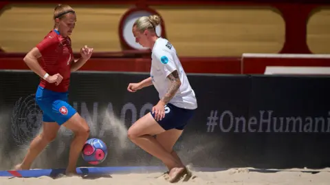 Jose Manuel Alvarez/Euro Beach Soccer A woman in a white kit runs towards a woman in a red and blue kit. Both women are looking at the ball in between them. They are on a beach.