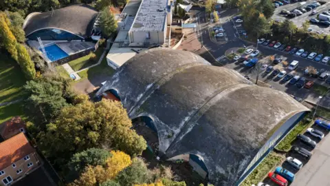 Historic England Archive Aerial view of aquatic centre, sports centre and former ice rink all with a thin reinforced concrete shell. Cars are parked to the north and east of the building and there is a border of trees at the rear, with a house the other side.