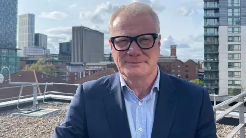 Richard Parker - a man with short white hair, glasses, a blue shirt and dark blue jacket, stands and smiles slightly. The view of a cityscape is visible behind him.