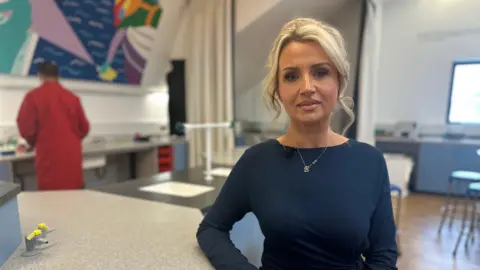 Abbie Yorke has blonde hair and is looking at the camera. She has a blue top and a silver necklace. She is sitting in a lab. 
