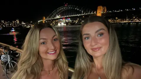 Fotografia de Charlie Buckland (à direita) e Chanel Buckland (à esquerda). Ambos têm olhos verdes e longos cabelos loiros e sorriem para a câmera. O fundo é escuro, mas atrás deles pode-se ver a Sydney Harbour Bridge, iluminando o rio abaixo. Atrás da ponte você pode ver arranha-céus, iluminados por fileiras de luz que entram pelas janelas. 