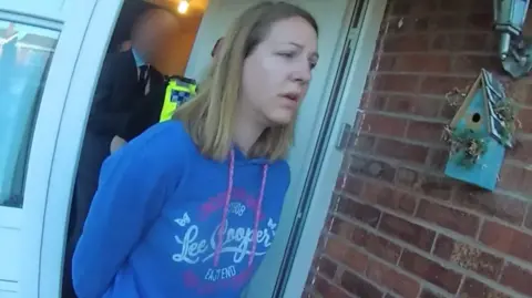 Cheshire Police Lucy Letby with long blond hair wearing a blue and white hoodie during her arrest. She has her arms behind her back and is walking out of a white front door with a police officer and a man in a navy suit whose face is blurred out behind her.