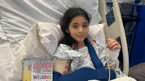 Young girl, Mina, in a hospital bed. Her arm is bandaged and in a sling. She has dark hair and smiles next to a 'get well soon' card.