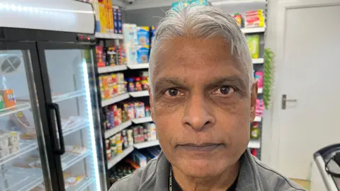 Bob Moodliar has short grey hair, with a small quiff at the top, he is wearing a grey fleece with a black shirt underneath, behind him are shelves of a grocery shop.