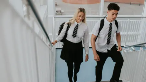 A girl (left) and a boy (right) walking up indoor school steps wearing school shirt and tie.