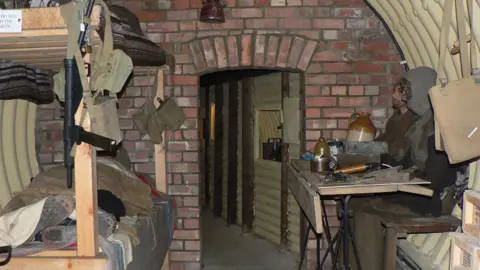 British Resistance Museum A replica of the Stratford St Andrew operating base. It has a curved metal roof and sides. There is a bunk bed on the left and a table with two mannequins sitting at it on the right. Beyond it is a brick wall leading to a corridor and an escape route.