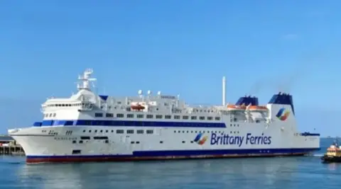 A white ferry with the Brittany Ferries logo on the side pulls into port on a calm, sunny day.