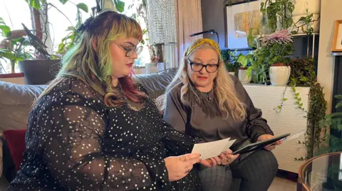 Daughter Charlie is wearing a black dress with stars and has green and pink dyed hair. Ian's widow Sarah is sat beside her on the settee, looking at old family photographs together.