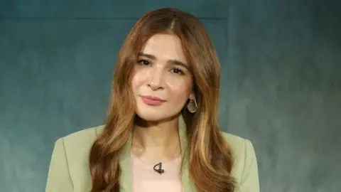 Headshot of a woman with long brown hair, looking into the camera. She is wearing a pale pink T-shirt under a beige jacket. She has a small microphone clipped to her top. 