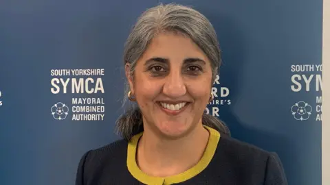 BBC A woman with grey hair is smiling and wearing a navy blue suit with yellow trim. She is stood in front of a blue board which says South Yorkshire SYMCA in white writing