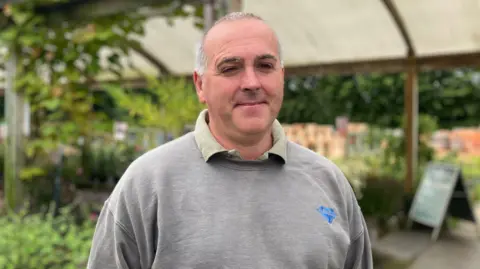 A man with short grey hair, wearing a grey jumper. There are plants behind him
