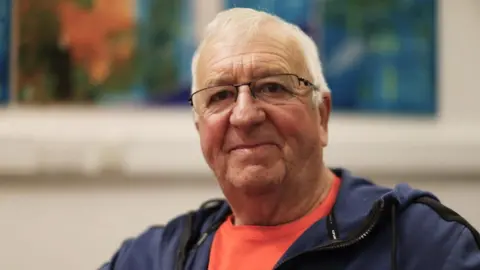 Harry Simpson, a man with white hair and glasses smiles at the camera. He is wearing an orange T shirt and a blue hooded top